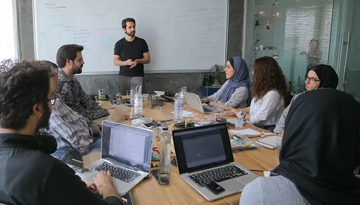 جلسه تیمی استارتاپی در اتاق کنفرانس، جایی که فردی در حال ارائه روی تخته سفید است و بقیه با لپتاپها مشغول یادداشتبرداری هستند.
