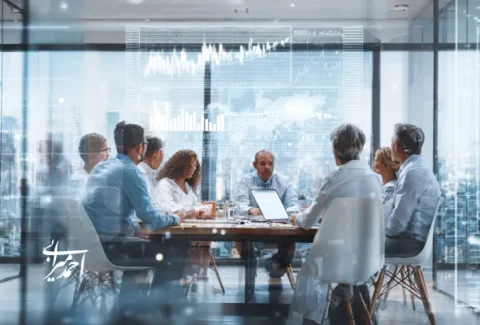 جلسه کاری گروهی در اتاق کنفرانس مدرن با دیوارهای شیشه‌ای، چند مدیر در حال بحث درباره نمودارهای مالی و استراتژی سرمایه‌گذاری، نمای شهری در پس‌زمینه.