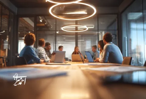جلسه تیمی در یک اتاق کنفرانس مدرن با طراحی صنعتی و نورپردازی حلقه‌ای، اعضای گروه در حال گفت‌وگو و کار با لپ‌تاپ‌ها دور میز جمع شده‌اند.