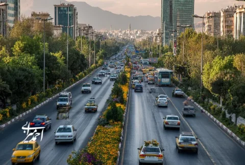 نمایی از بزرگراه شلوغ در تهران با درختان سبز و کوه‌های البرز در پس‌زمینه.