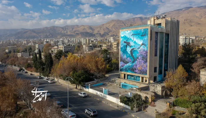 نمایی از خیابانی در شهر با ساختمانی بزرگ که روی دیوارش نقاشی هنری رنگارنگی از پرندگان و طبیعت دیده می‌شود.