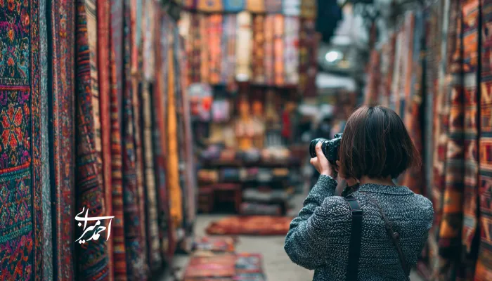 یک گردشگر در حال عکاسی از بازار سنتی پر از فرش‌ها و قالی‌های رنگارنگ ایرانی؛ نمایی از هویت فرهنگی، صنایع‌دستی و زیبایی بصری بازارهای بومی ایران