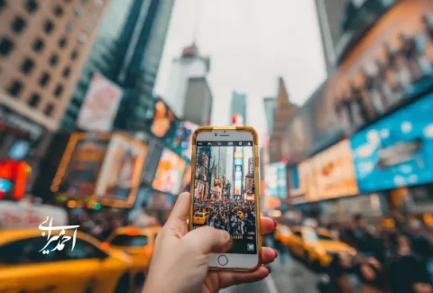 دست فردی که با گوشی هوشمند از خیابان شلوغی با تاکسی‌های زرد و بیلبوردهای رنگارنگ در مرکز شهر عکس می‌گیرد؛ تصویری از تجربه شهری پرانرژی و برندینگ گردشگری مدرن.