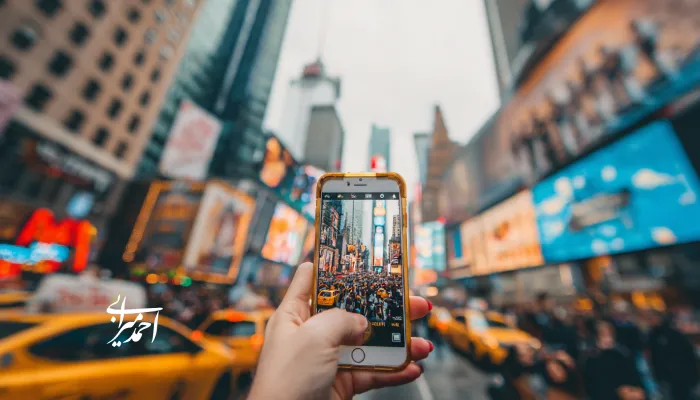 دست فردی که با گوشی هوشمند از خیابان شلوغی با تاکسی‌های زرد و بیلبوردهای رنگارنگ در مرکز شهر عکس می‌گیرد؛ تصویری از تجربه شهری پرانرژی و برندینگ گردشگری مدرن.