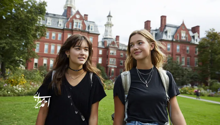 دو دختر نوجوان در محوطه دانشگاه هاروارد در حال قدم‌زدن هستند؛ با کوله‌پشتی، لباس‌های ساده و چهره‌ای آرام و خندان، در میان ساختمان‌های آجری و فضای سبز دانشگاه.