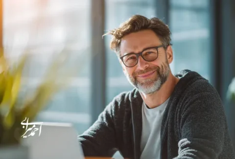 مدیر میانسال با عینک و لبخند آرام، پشت میز کار در یک دفتر روشن و مدرن نشسته و به دوربین نگاه می‌کند؛ نور طبیعی از پنجره و گیاه آپارتمانی در کنار او دیده می‌شود.