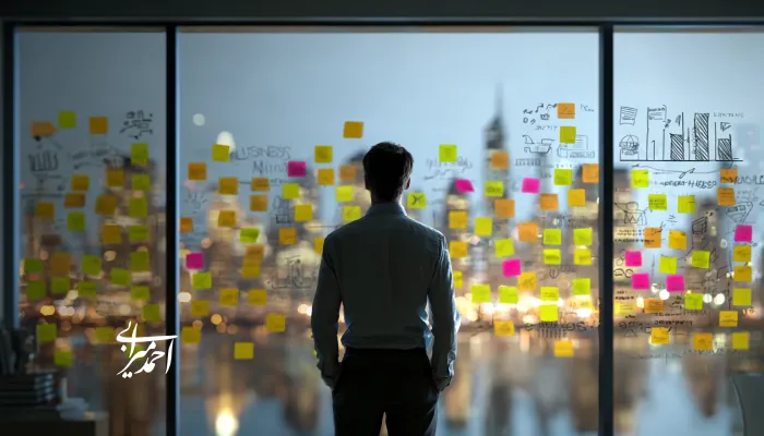 مدیری از پشت‌سر مقابل شیشه‌ای پر از یادداشت‌های چسبان و طرح‌های نمودار ایستاده و به منظره شهر در پس‌زمینه نگاه می‌کند؛ فضایی مرتبط با برنامه‌ریزی، تصمیم‌گیری و استراتژی کسب‌وکار.