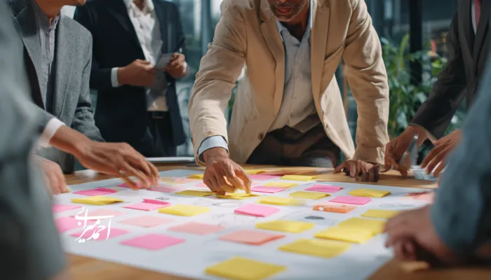 کارگاه هم‌فکری تیمی در فضای کاری؛ چند نفر با لباس رسمی دور یک میز ایستاده‌اند و روی یک پوستر بزرگ، یادداشت‌های چسبی رنگی را برای برنامه‌ریزی و ایده‌پردازی جابه‌جا می‌کنند.