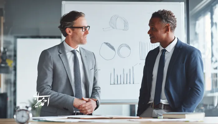 دو مدیر در حال گفت‌وگوی کوتاه و حرفه‌ای در دفتر کار کنار وایت‌برد با نمودارها و یک تایمر رومیزی، نمادی از مدیریت یک‌دقیقه‌ای و بازخورد سریع.